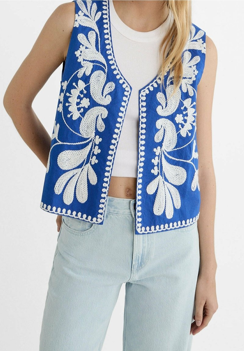Femme portant un gilet bleu sans manches avec une broderie florale blanche sur un crop top blanc et un jean bleu clair.