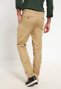 Chinos couleur fauve à coupe slim, comprenant des poches arrière avec des coutures discrètes et une texture lisse, assortis à des baskets noires.