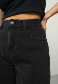 Svarta denimjeans med hög midja, framfickor och en liten silverknapp vid midjan, med synliga sömmar.