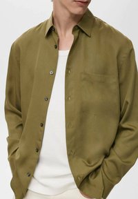 Olive green button-up shirt made of smooth fabric, featuring a chest pocket, pointed collar, and black buttons. Worn over a white T-shirt.