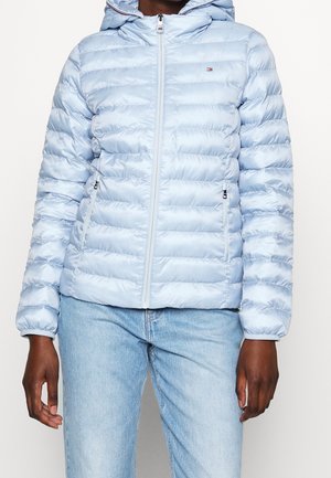 Person trägt eine hellblaue gesteppte Kapuzenjacke mit Frontreißverschluss und Reißverschlusstaschen, kombiniert mit hellblauen Jeans.