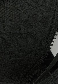 Tissu noir texturé avec un motif tissé, présentant des détails élaborés et un bord festonné. Comprend des sangles élastiques douces pour un ajustement.