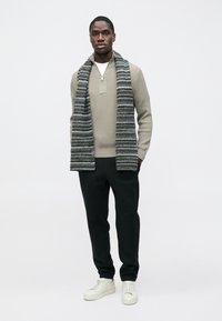 Pull beige clair zippé avec texture côtelée, pantalon noir et écharpe grise rayée. Le mannequin portant des baskets blanches se tient devant un fond uni.