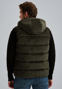 Homme portant un gilet à capuche matelassé vert foncé sur un t-shirt noir à manches longues et un jean bleu, vu de dos contre un fond uni.