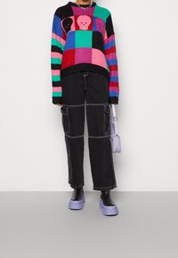 Maglione lavorato a maglia colorato con strisce nere, rosa, verdi e blu; presenta facce grafiche. Pantaloni cargo neri; scarpe con zeppa viola; piccola borsa viola.