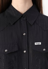 Black button-up shirt with a textured fabric, vertical stripes, and two front pockets. Features a collar and branded label on the lower side.