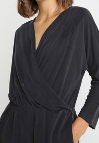 Robe enveloppante noire à manches longues en tissu doux et texturé. Dispose d'un décolleté en V et d'une taille froncée pour une silhouette fluide.