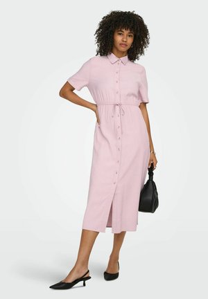Kobieta o kręconych włosach ubrana w jasnoróżową sukienkę midi na guziki z wiązanym paskiem w talii, trzyma czarną torebkę i nosi czarne szpilki.