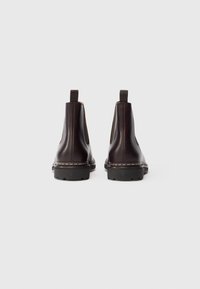 Une paire de bottes Chelsea marron foncé avec des panneaux élastiques noirs sur les côtés et des semelles épaisses noires, vues de l'arrière sur un fond blanc.
