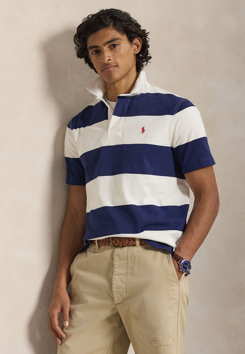 Polo à rayures bleu marine et blanc avec un col, arborant un petit logo rouge. Porté avec un pantalon beige et une ceinture tressée marron.