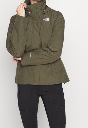 Persoon draagt een olijfgroene North Face jas met een hoge kraag en zwarte broek, houdt de kraag met één hand vast terwijl hij tegen een witte achtergrond staat.