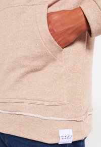 Béžový fleecový svetr s přední kapsou, šitým lemem a štítkem dole vlevo. Tkanina má strukturovaný, měkký vzhled.