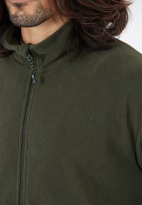 Chaqueta de forro polar verde con un alto cuello y cierre de cremallera. El material texturizado presenta un logo sutil del mismo color en el pecho.