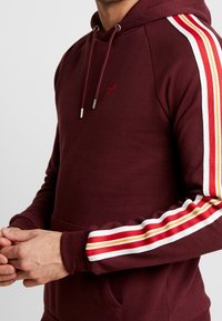 Sudadera burdeos con bolsillo frontal y capucha con cordón. Presenta acentos en blanco y rayas rojas, amarillas y naranjas a lo largo de las mangas.