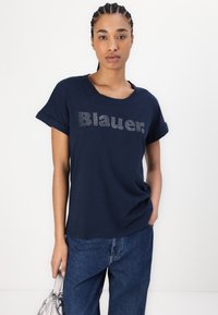 Mørkeblå bomull t-skjorte med oppbrettede ermer, med en rhinestone-prydet "Blauer." logo. Brukt med vide denimbukser.