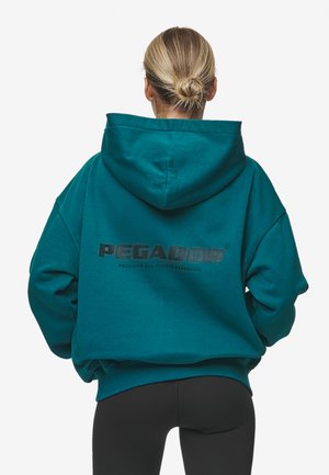 Personne aux cheveux blonds en chignon portant un sweat à capuche bleu sarcelle avec le texte « PEGADOR » dans le dos et des leggings noirs, debout de dos.