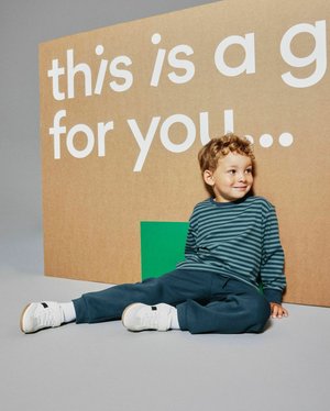 Młody chłopiec w pasiastym, długim rękawie koszuli i ciemnych spodniach siedzi na podłodze, opierając się o oznakowanie z częściowym białym tekstem na brązowym tle.