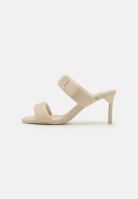 Beige hæler sandaler med en glat imitation læder finish, der har to brede stropper og en slank høj hæl. Diskret logo detalje på stroppen.