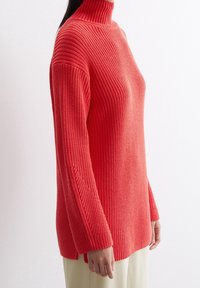 Roter Strickpullover mit Rollkragen, gerippter Textur, überschnittenen Schultern und geteiltem Saum. Kombiniert mit cremefarbenen Hosen, die die Ärmellänge hervorheben.