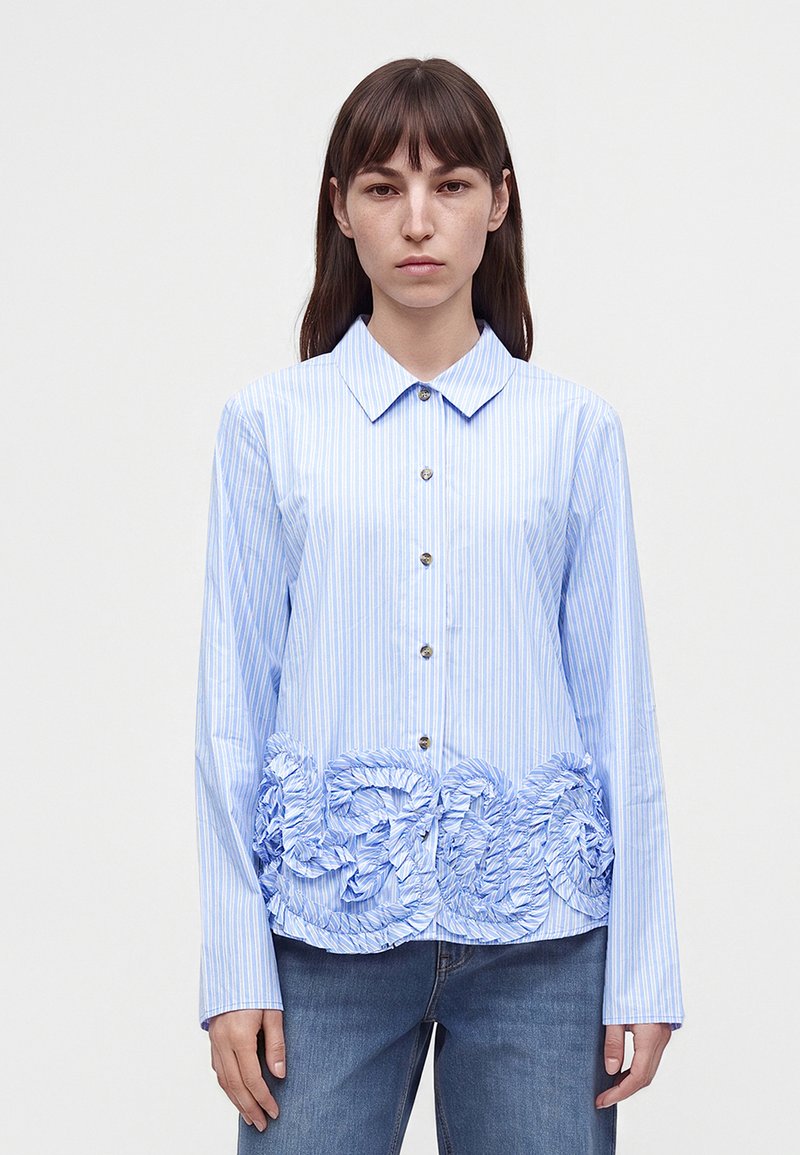 Femme portant une chemise rayée bleu clair à boutons avec un détail en tissu texturé et froncé au niveau de la taille, associée à un jean bleu.