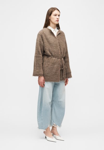 Γυναίκα που φοράει καφέ χνουδωτό σακάκι με ζώνη, λευκό πουκάμισο, ανοιχτό μπλε τζιν με φαρδιά μπατζάκια και frayed άκρες, και λευκά ψηλοτάκουνα παπούτσια με μυτερή μύτη, όρθια μπροστά σε λευκό φόντο.