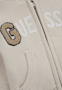 Pull beige à fermeture éclair, avec un tricot texturé, arborant le logo brodé "GUESS" en beige et blanc, et deux poches latérales.