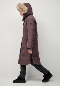 Lang, lilla parka med en hætte i pels, frontlynlås og sidelommer. Stof ser glat ud, parret med sorte casual sko.