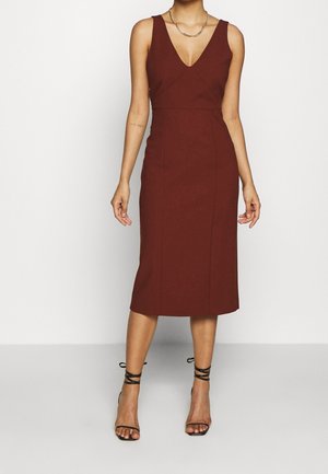 Persona que lleva un vestido midi burdeos sin mangas y ajustado, con escote en V y sandalias negras de tacón con tiras, de pie sobre un fondo liso.