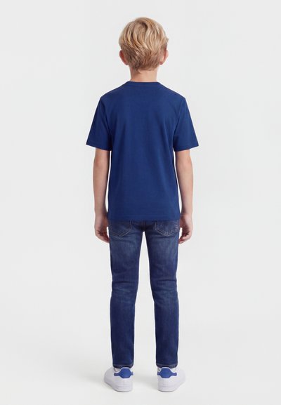 Ξανθό αγόρι που στέκεται με την πλάτη, φοράει μπλε σκούρο μπλουζάκι, σκούρο μπλε τζιν και άσπρα αθλητικά με μπλε λεπτομέρειες.