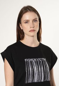 Camiseta de algodón negra con mangas cortas, que presenta un texto bordado en blanco que dice "FASHION VICTIM" y un detalle de flecos largos debajo del texto.