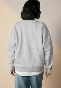 Persona con capelli corti e scuri che indossa una felpa grigia chiara e jeans blu, in piedi di fronte a uno sfondo beige semplice.