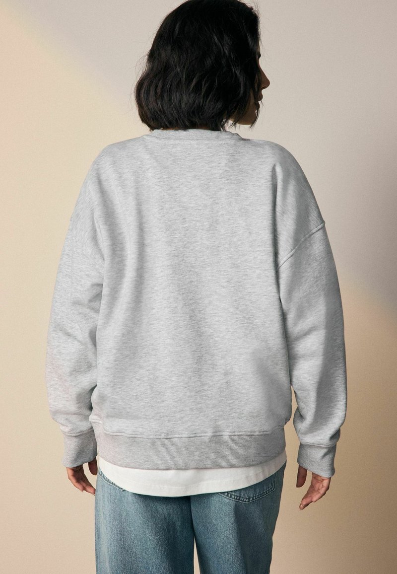 Persona con capelli corti e scuri che indossa una felpa grigia chiara e jeans blu, in piedi di fronte a uno sfondo beige semplice.
