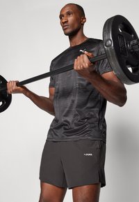 Zwarte sport t-shirt met subtiel patroon, voorzien van een logo. Gecombineerd met grijze shorts, die een lichtgewicht ontwerp tonen. Gewichten vastgehouden in de handen.