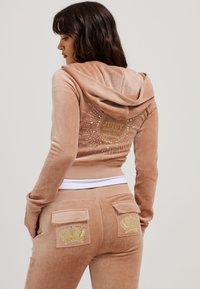 Lichtbruine velours hoodie met een capuchon en een achterdesign met gouden branding en rhinestones. Gecombineerd met bijpassende broek met kroonaccenten.