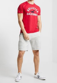 Punainen puuvillapaita, jossa on valkoisella painettu "HOLLISTER CALIFORNIA". Yhdistetty harmaan heather-kankaan shortseihin ja valkoisiin lenkkareihin, joissa on mustia yksityiskohtia.