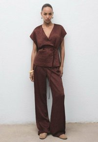 Burgundy wrap-top dengan lengan pendek dan kain bertekstur, dipadukan dengan celana lebar dengan warna yang sama. Aksesori termasuk gelang besar.
