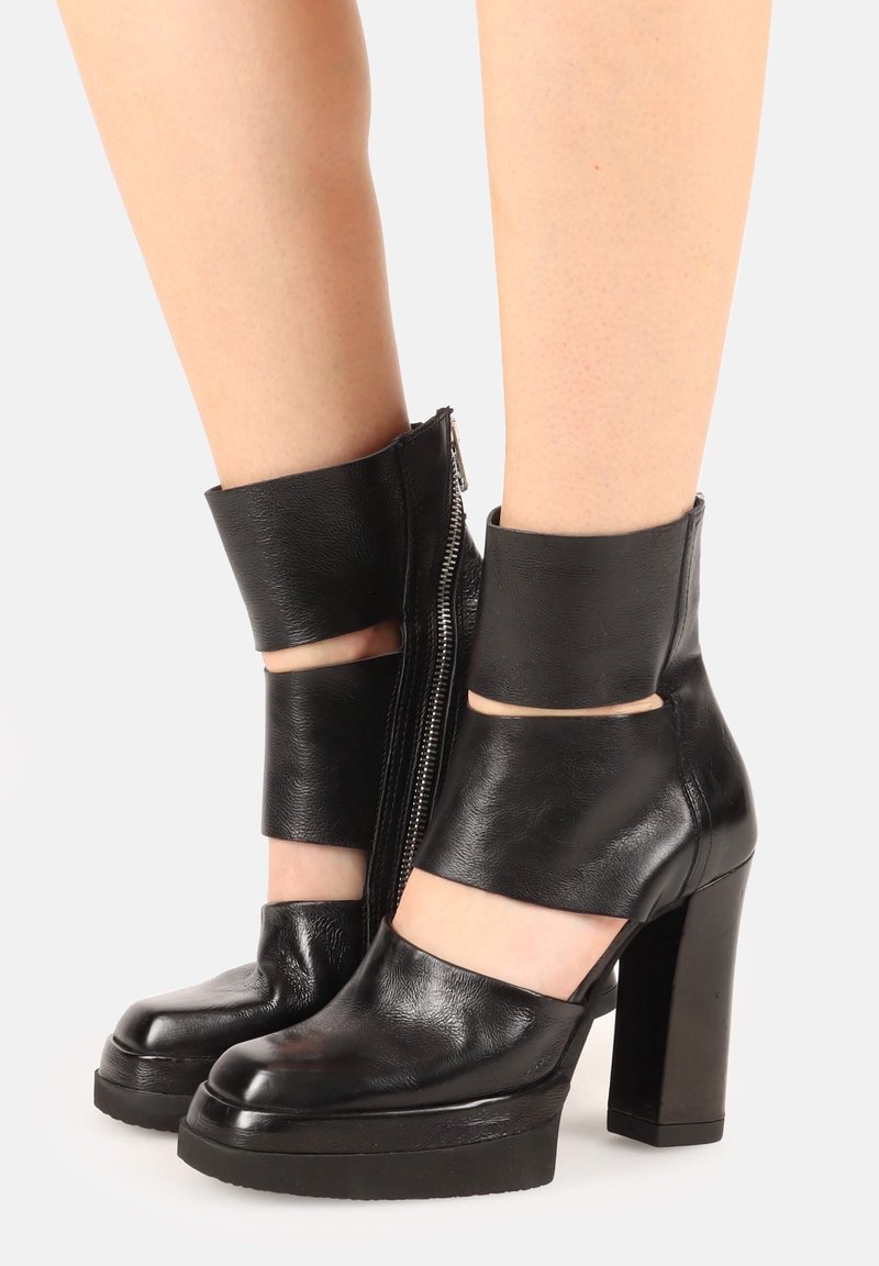 Black leather ankle boots with a block heel, featuring cut-outs and an ankle strap, highlighted by a side zipper for closure.