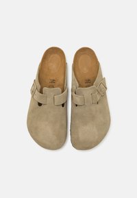 Beige semsketøfler med åpen bak, rund tå og en dekorativ spenne. Har en teksturert overdel og glatt lærsåle.