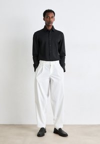 Black, long-sleeve shirt with buttons, paired with white, pleated trousers; black slip-on shoes complete the outfit. Smooth fabric and structured fit.