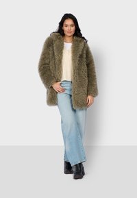 Green faux fur coat with a wide collar, worn over a cream sweater and light blue high-waisted jeans. Model stands with hands on hips.