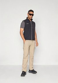 Juoda be rankovių liemenė dėvima ant pilkos trumpomis rankovėmis marškinėlių, smėlio spalvos krovinių tipo kelnės ir juodi batai; priderinti tamsūs saulės akiniai.