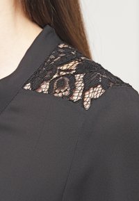 Blusa nera in tessuto morbido con un delicato accento in pizzo sulle spalle, che presenta motivi floreali e un colletto strutturato.
