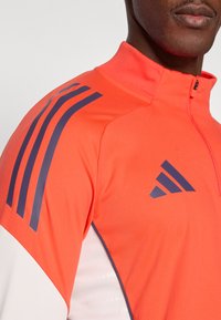 Oransje treningsjakke med høy krage, marineblå striper på skulderen og en logo. Laget av glatt, elastisk stoff med glidelås foran.