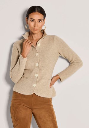 Frau in beiger Knopfpullover-Strickjacke mit Stehkragen und braunen Wildlederhosen, die mit einer Hand auf der Hüfte und der anderen nah am Kragen posiert.