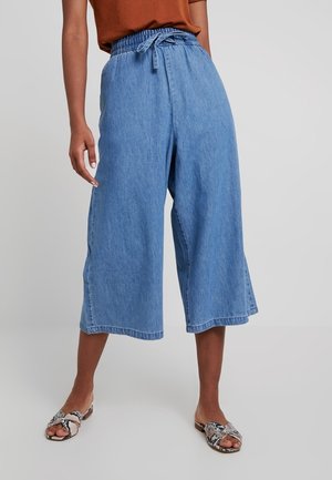 Kvinde iført blå denim vidde-cropped bukser med snøre i taljen og åbne flade sandaler med slangeprint.