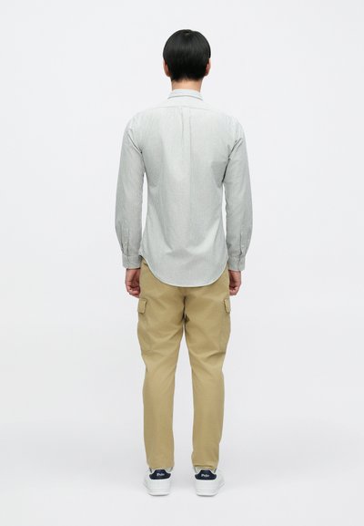 Pánská košile s dlouhým rukávem a svislými šedými pruhy, doplněná béžovými kalhotami typu cargo a bílými teniskami, pohled zezadu.