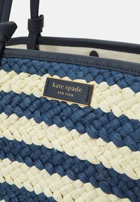 Woven tote bag featuring dark blue and cream stripes, crafted from straw with black leather accents and a gold logo plate.
