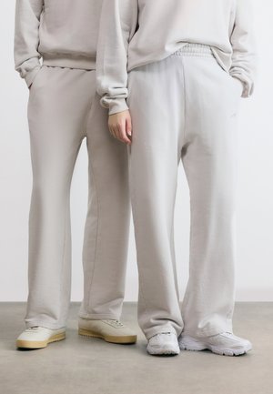 Δύο άτομα φορούν μπεζ φόρμες και ανοιχτόχρωμα αθλητικά παπούτσια, στέκονται δίπλα-δίπλα με τα χέρια στις τσέπες, μπροστά σε ένα απλό φόντο.