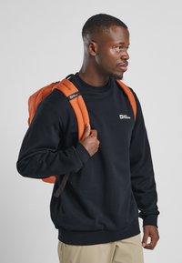 Sweatshirt noir à col rond porté sur un pantalon kaki, associé à un sac à dos orange avec des bretelles rembourrées et un détail de logo.