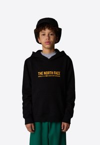 Felpa con cappuccio nera e tasca frontale; logo "THE NORTH FACE" in giallo. Indossata con pantaloni corti verdi e un cappello a secchiello nero.